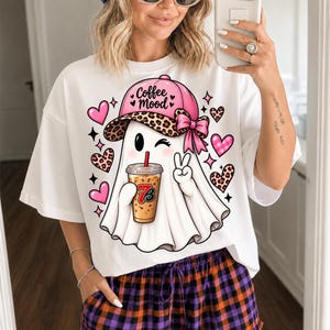 May include: A white t-shirt featuring a graphic of a ghost wearing a pink leopard print baseball cap with a bow. The ghost is winking, holding an iced coffee with a straw, and making a peace sign. The design includes pink and leopard print hearts.