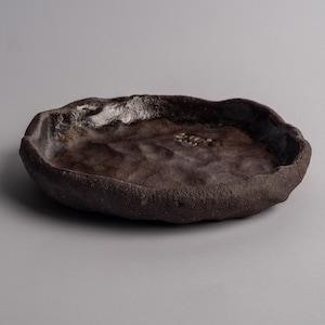 May include: A shallow, circular ceramic dish with a dark brown, textured exterior and a glossy, uneven interior surface. The rim is thick and irregular, featuring a hand-formed appearance with a deep, dark brown finish throughout the piece.