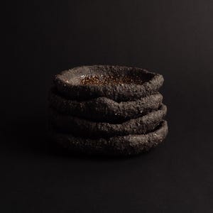 May include: A stack of four circular, shallow bowls with a dark, coarse, and heavily textured exterior surface. The interior of the top bowl features a glossy, speckled brown finish that contrasts with the matte, black, rough-hewn outer edges.