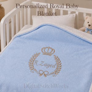 Peut inclure: Une couverture pour bébé bleu clair avec une doublure en sherpa blanc. Le centre présente une broderie dorée d'une couronne au-dessus d'une couronne de laurier entourant le texte R. Zayed. Le texte Personalized Royal Baby Blanket est visible.