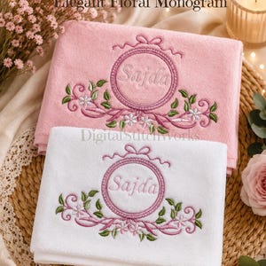 Peut inclure: Deux serviettes brodées, une rose et une blanche, avec un motif de monogramme floral. Les serviettes présentent un cadre circulaire avec le nom "Sajda" brodé dans une couleur assortie, entouré de détails floraux et de rubans. Le texte "Elegant Floral Monogram" est en haut.