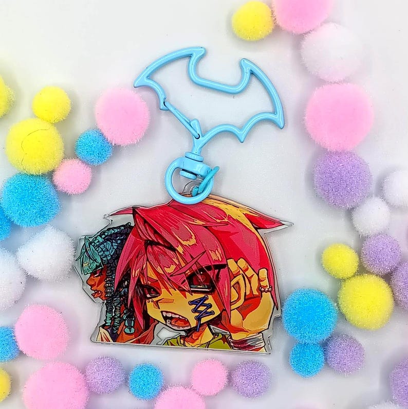 May include: A light blue acrylic keyring featuring a bat-shaped clasp and a printed illustration of a character with bright red hair, sharp features, and a blue stitched mark on their cheek, positioned alongside another character with blue hair.