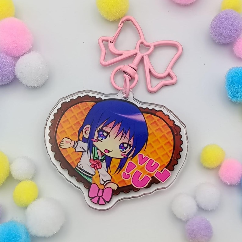 May include: A pink bow-shaped keychain clip attached to a clear acrylic charm. The charm features a cartoon anime character with blue hair, a school uniform, and the text "VU!" on a heart-shaped waffle background.