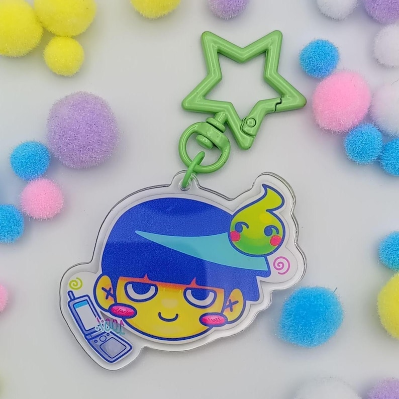 May include: A clear acrylic keychain with a cartoon character design. The character has blue hair, a yellow face, and a green accent. The keychain has a green star-shaped clasp and a small flip phone graphic. Colourful pom-poms surround the design.