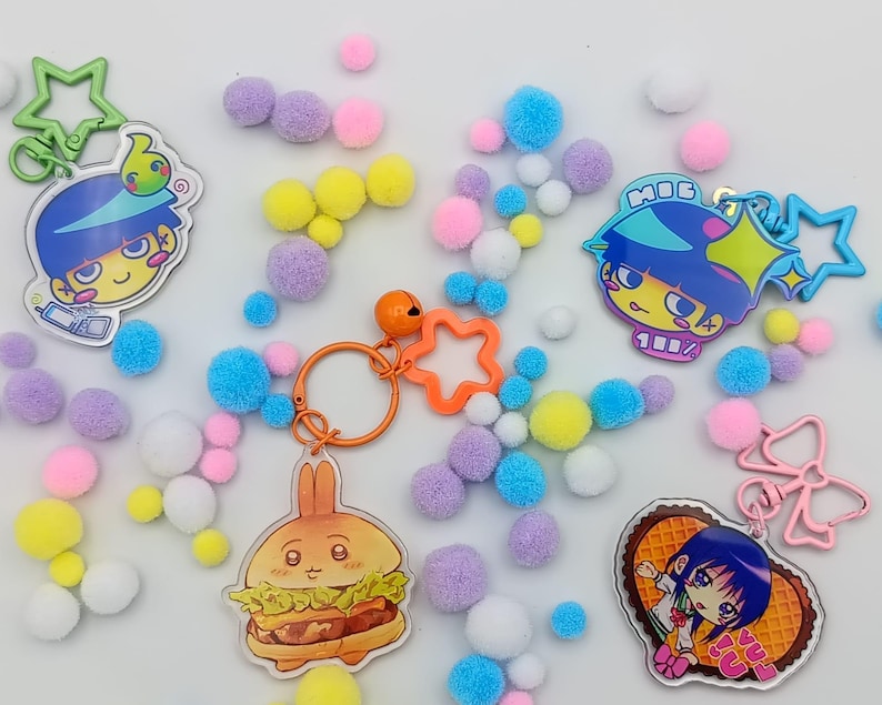 May include: A selection of vibrant keyrings with cartoon characters and food designs. The keyrings include star, bow, and burger shapes. Scattered around are small, fluffy pom-poms in pink, yellow, blue, and purple hues.