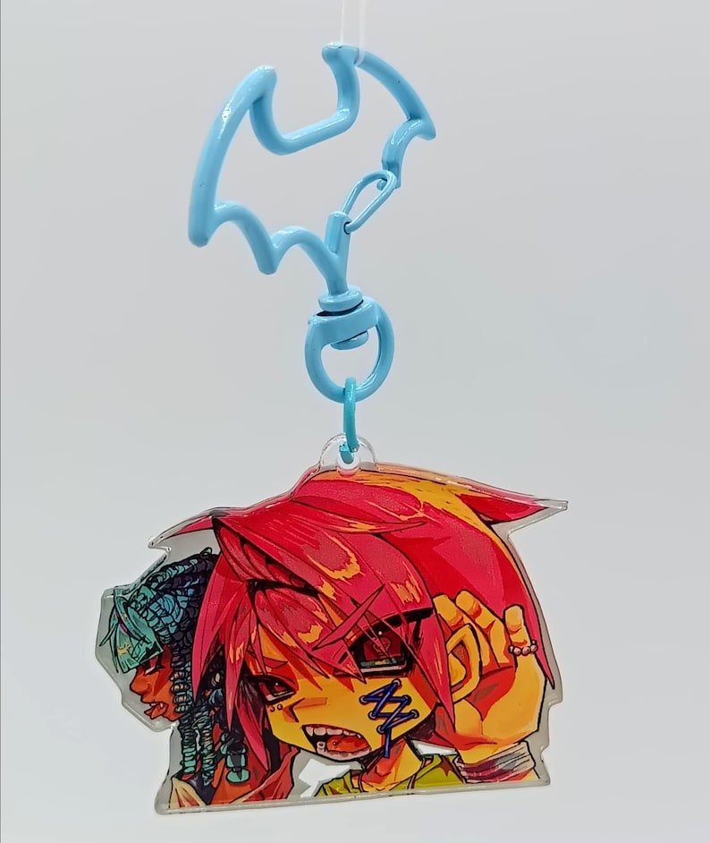 May include: A clear acrylic keyring featuring a light blue bat-shaped clasp. The charm displays a colourful illustration of two characters with stylised hair, one with vibrant pink hair and a blue stitched mark on the cheek, and another with teal hair.