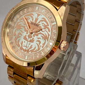 May include: A gold-toned Guess watch with a lion design on the face. The watch features a gold band and a round face with the brand name and a lion graphic. The watch is displayed on a clear stand.