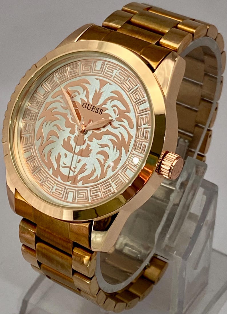 May include: A gold-toned Guess watch with a lion design on the face. The watch features a gold band and a round face with the brand name and a lion graphic. The watch is displayed on a clear stand.