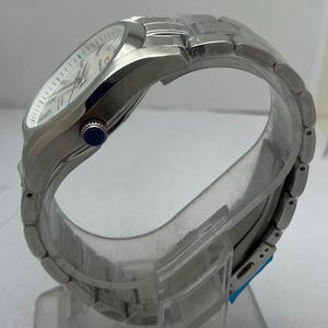May include: A silver-toned stainless steel wristwatch featuring a white dial with silver indices and hands. The watch has a metallic link bracelet and a side crown with a blue circular accent featuring the letters GS in white text.