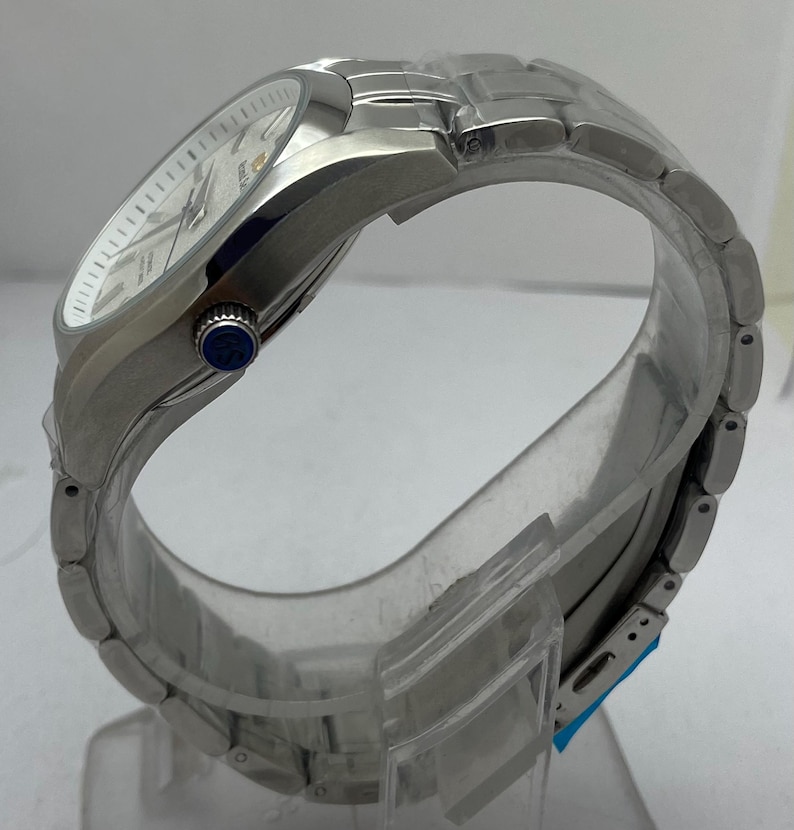 May include: A silver-toned stainless steel wristwatch featuring a white dial with silver indices and hands. The watch has a metallic link bracelet and a side crown with a blue circular accent featuring the letters GS in white text.