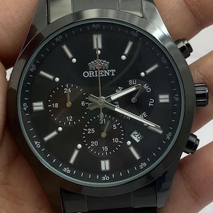 May include: A black stainless steel Orient chronograph watch with a matching link bracelet. The black dial features luminous hour markers, three sub-dials, a date window at the four o'clock position, and the Orient logo printed in white at the top.
