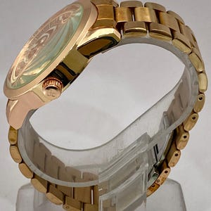 May include: A gold-toned wristwatch with a round face and a matching link bracelet. The watch face has a light-colored, patterned design. The watch is displayed on a clear stand, showcasing its side profile and the details of the band.