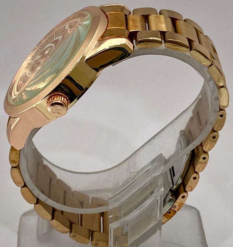 May include: A gold-toned wristwatch with a round face and a matching link bracelet. The watch face has a light-colored, patterned design. The watch is displayed on a clear stand, showcasing its side profile and the details of the band.