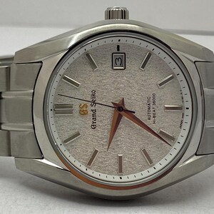 May include: A silver-toned Grand Seiko wristwatch with a textured light-coloured dial, silver hour markers, and a date window at the three o'clock position. The watch features a metallic link bracelet and gold-toned hands with a gold GS logo.
