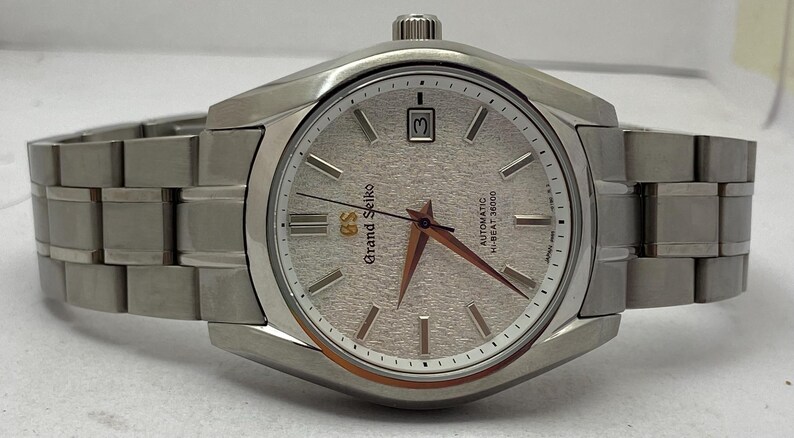 May include: A silver-toned Grand Seiko wristwatch with a textured light-coloured dial, silver hour markers, and a date window at the three o'clock position. The watch features a metallic link bracelet and gold-toned hands with a gold GS logo.