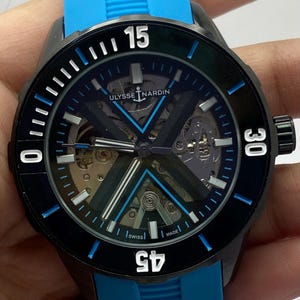 May include: A Ulysse Nardin wristwatch with a black case, a skeletonized dial featuring a prominent X-shaped bridge, and a bright blue rubber strap. The black bezel displays white numerals 15, 30, 45, and 0, with blue accents on the indices.