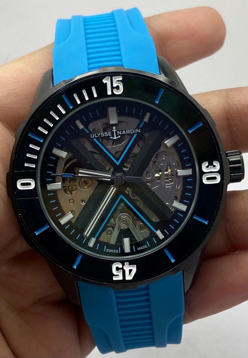 May include: A Ulysse Nardin wristwatch with a black case, a skeletonized dial featuring a prominent X-shaped bridge, and a bright blue rubber strap. The black bezel displays white numerals 15, 30, 45, and 0, with blue accents on the indices.