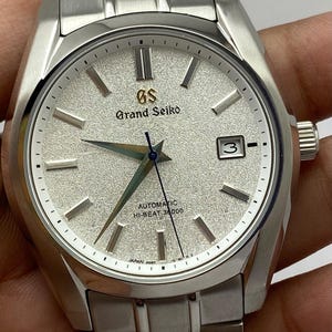 May include: A silver-toned Grand Seiko wristwatch featuring a textured white dial, silver hour markers, and a date window at the three o'clock position. The watch includes a stainless steel link bracelet, a blue second hand, and the text GS Grand Seiko Automatic Hi-Beat 36000.