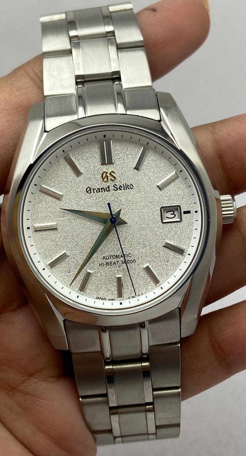 May include: A silver-toned Grand Seiko wristwatch featuring a textured white dial, silver hour markers, and a date window at the three o'clock position. The watch includes a stainless steel link bracelet, a blue second hand, and the text GS Grand Seiko Automatic Hi-Beat 36000.