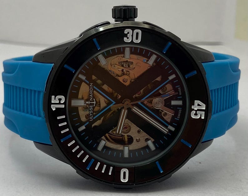May include: A Ulysse Nardin wristwatch featuring a black case and a bright blue rubber strap. The dial is skeletonized, revealing internal gold-toned mechanical components, with a black X-shaped bridge, white hour markers, and blue accents.
