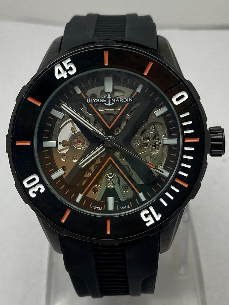 K&ouml;nnte beinhalten: Eine schwarze Ulysse Nardin Armbanduhr mit skelettiertem Zifferblatt, X-f&ouml;rmiger Br&uuml;cke, orangefarbenen Akzenten und wei&szlig;en Leuchtmarkierungen. Die Uhr verf&uuml;gt &uuml;ber eine schwarze Drehl&uuml;nette mit wei&szlig;en Ziffern und ein schwarzes Kautschukarmband.