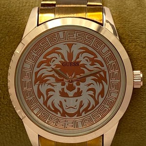 May include: A gold-toned Guess wristwatch with a round face featuring a lion design. The watch has a gold band and a textured bezel. The watch face displays the Guess logo and is set against a light background.