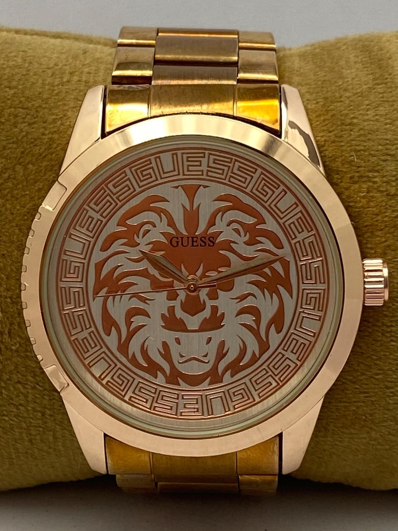 May include: A gold-toned Guess wristwatch with a round face featuring a lion design. The watch has a gold band and a textured bezel. The watch face displays the Guess logo and is set against a light background.