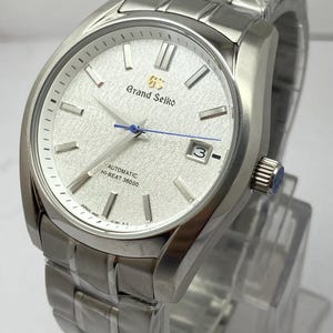 May include: A silver-tone Grand Seiko automatic watch with a textured white dial, silver indices, a blue second hand, and a date window at the three o'clock position. The watch features a stainless steel link bracelet and a crown with a logo.