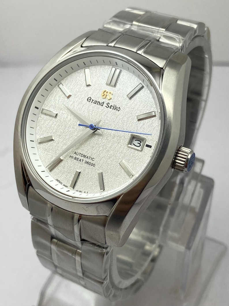 May include: A silver-tone Grand Seiko automatic watch with a textured white dial, silver indices, a blue second hand, and a date window at the three o'clock position. The watch features a stainless steel link bracelet and a crown with a logo.