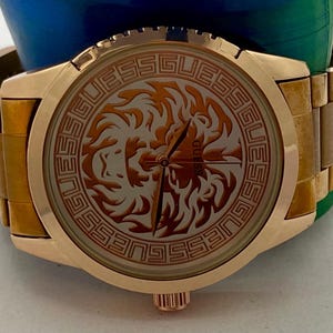 May include: Gold-toned Guess watch with a round face featuring a lion design. The watch has a gold band and a gold bezel. The watch face has the word "GUESS" around the lion design.