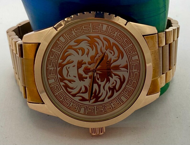 May include: Gold-toned Guess watch with a round face featuring a lion design. The watch has a gold band and a gold bezel. The watch face has the word "GUESS" around the lion design.