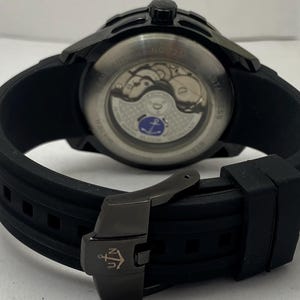 K&ouml;nnte beinhalten: Eine schwarze Armbanduhr mit Silikonarmband und einer schwarzen Metallschnalle mit Anker-Logo. Der Geh&auml;useboden ist transparent und gibt den Blick auf das mechanische Uhrwerk sowie ein kleines blaues Anker-Emblem auf dem Rotor frei.