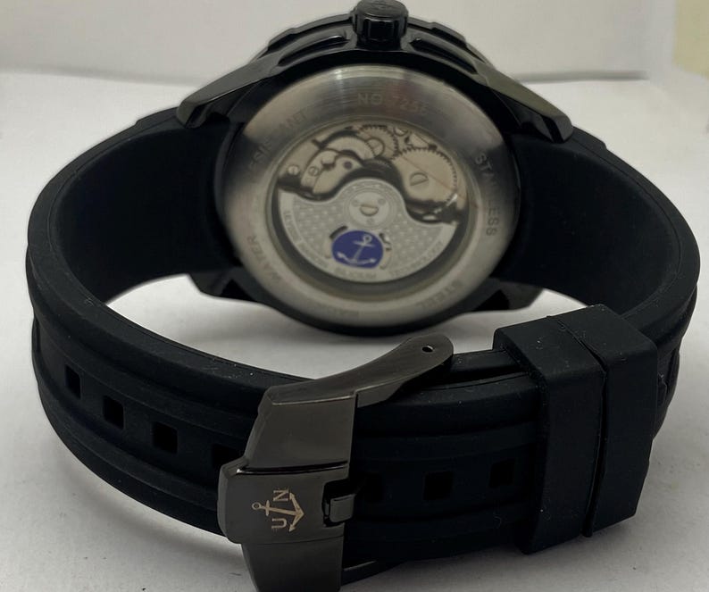 K&ouml;nnte beinhalten: Eine schwarze Armbanduhr mit Silikonarmband und einer schwarzen Metallschnalle mit Anker-Logo. Der Geh&auml;useboden ist transparent und gibt den Blick auf das mechanische Uhrwerk sowie ein kleines blaues Anker-Emblem auf dem Rotor frei.