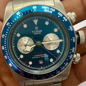 May include: A stainless steel Tudor Geneve chronograph watch with a blue dial and a blue tachymeter bezel. The watch features two silver sub-dials, a date window at the six o'clock position, and a metal link bracelet. The text reads Tudor Geneve.