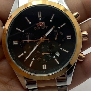 May include: An Orient chronograph wristwatch featuring a black dial with rose gold-tone hands and hour markers. The watch has a two-tone stainless steel bracelet in silver and rose gold, a gold-tone bezel, and three sub-dials with a date window.