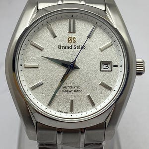 May include: A silver-toned stainless steel wristwatch featuring a textured white dial, silver hour markers, and a date window at the three o'clock position. The dial displays the Grand Seiko logo, Automatic, and Hi-Beat 36000 text in black.