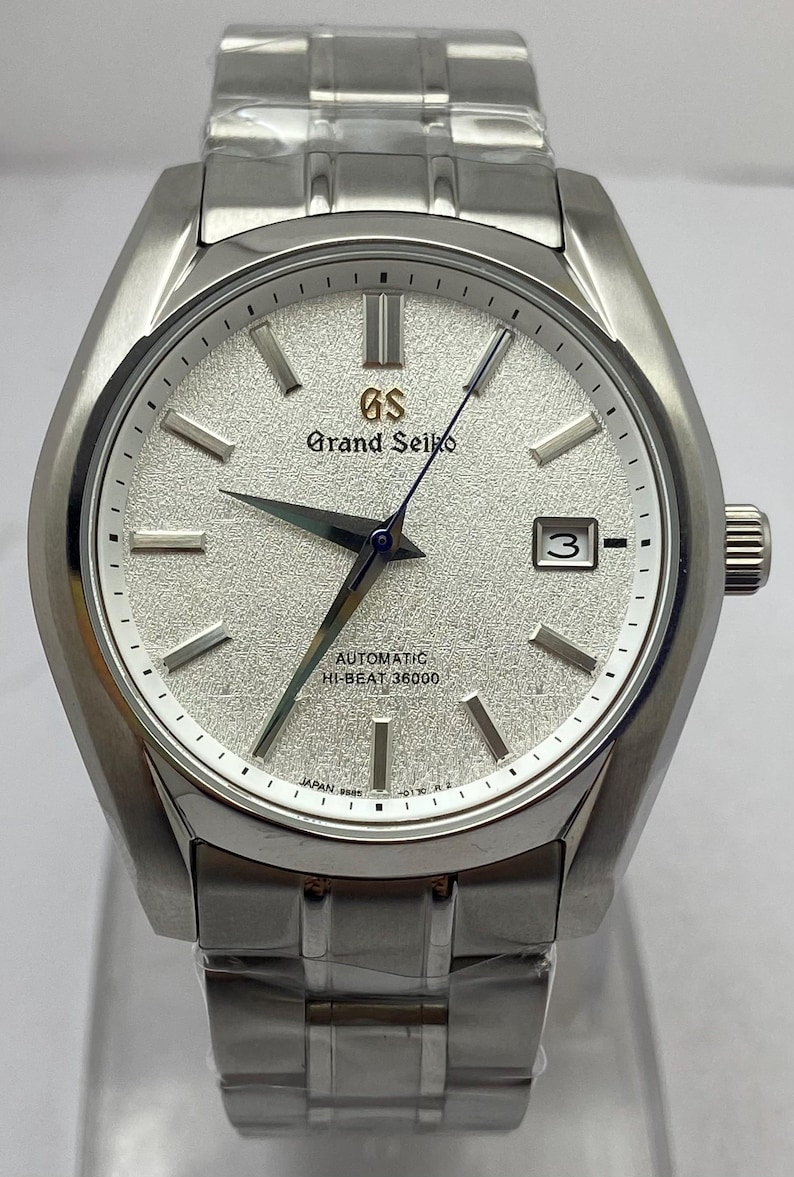 May include: A silver-toned stainless steel wristwatch featuring a textured white dial, silver hour markers, and a date window at the three o'clock position. The dial displays the Grand Seiko logo, Automatic, and Hi-Beat 36000 text in black.