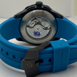 May include: A wristwatch with a bright blue silicone strap and a black buckle featuring an anchor logo. The watch case is black with a transparent exhibition case back revealing the internal mechanical movement and a small blue anchor emblem.