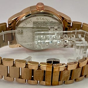 May include: Rose gold-toned wristwatch with a stainless steel back and a matching link bracelet. The watch is displayed on a clear plastic stand. The watch face is visible, and the back of the watch has the text "STAINLESS".