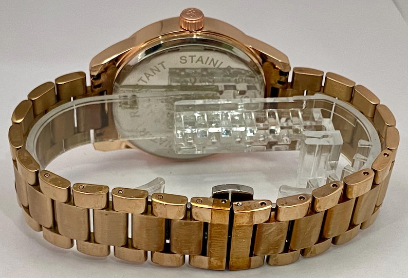 May include: Rose gold-toned wristwatch with a stainless steel back and a matching link bracelet. The watch is displayed on a clear plastic stand. The watch face is visible, and the back of the watch has the text "STAINLESS".