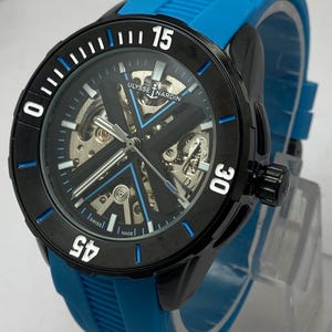 May include: A Ulysse Nardin wristwatch with a black circular bezel featuring white numerals 15, 30, and 45. The watch has a skeletonized dial with a prominent black X-shaped bridge, blue accents, and a bright blue textured rubber strap.