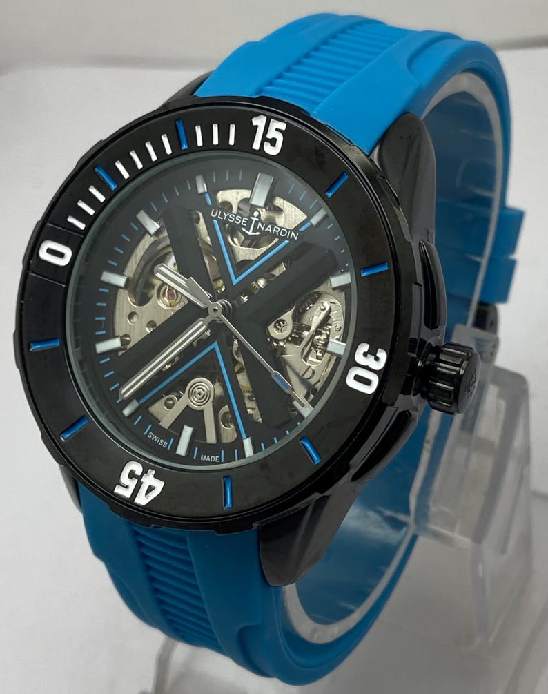 May include: A Ulysse Nardin wristwatch with a black circular bezel featuring white numerals 15, 30, and 45. The watch has a skeletonized dial with a prominent black X-shaped bridge, blue accents, and a bright blue textured rubber strap.