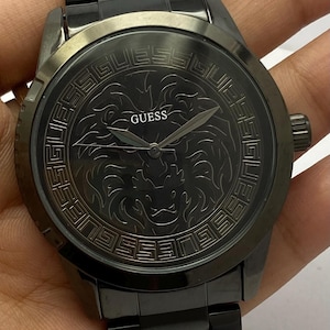 May include: A black Guess watch with a matching black strap. The watch face features a lion design and the Guess logo. The watch has a dark, sleek appearance.