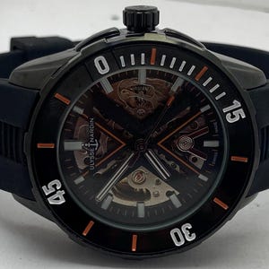 K&ouml;nnte beinhalten: Eine schwarze Ulysse Nardin Armbanduhr mit skelettiertem Zifferblatt und orangefarbenen sowie wei&szlig;en Stundenmarkierungen auf der L&uuml;nette. Die Uhr hat ein schwarzes Kautschukarmband und ein schwarzes Geh&auml;use mit einer Krone oben. Der Markenname ist sichtbar.