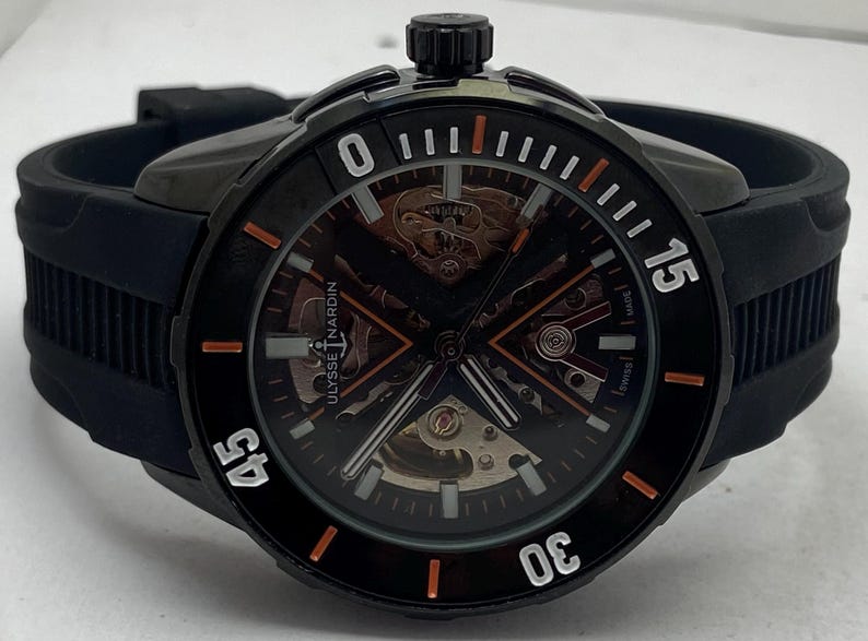 K&ouml;nnte beinhalten: Eine schwarze Ulysse Nardin Armbanduhr mit skelettiertem Zifferblatt und orangefarbenen sowie wei&szlig;en Stundenmarkierungen auf der L&uuml;nette. Die Uhr hat ein schwarzes Kautschukarmband und ein schwarzes Geh&auml;use mit einer Krone oben. Der Markenname ist sichtbar.