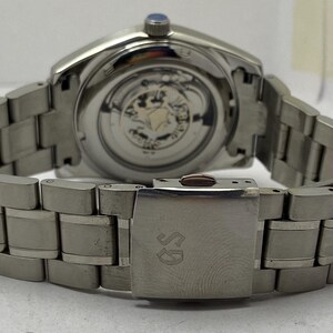 May include: A silver-toned stainless steel wristwatch featuring a metallic link bracelet with a deployment clasp engraved with the letters GS. The watch case back is transparent, revealing the internal mechanical movement and gears inside.