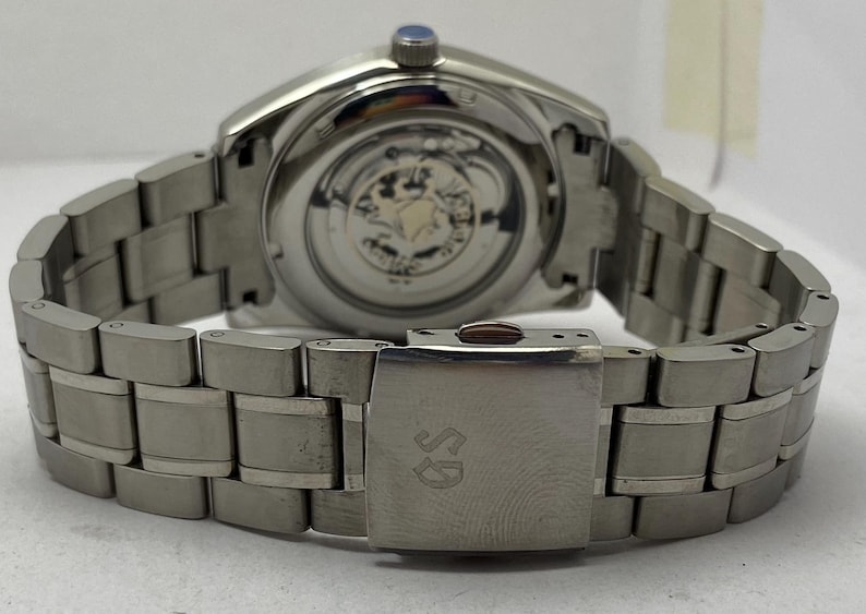 May include: A silver-toned stainless steel wristwatch featuring a metallic link bracelet with a deployment clasp engraved with the letters GS. The watch case back is transparent, revealing the internal mechanical movement and gears inside.