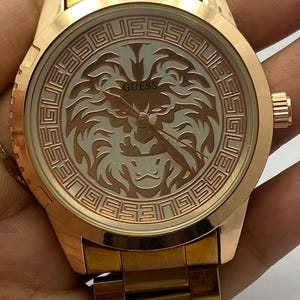 May include: Rose gold-toned Guess watch with a round face. The watch face features a lion design and the Guess logo. The watch has a matching rose gold band. The watch is a stylish accessory.