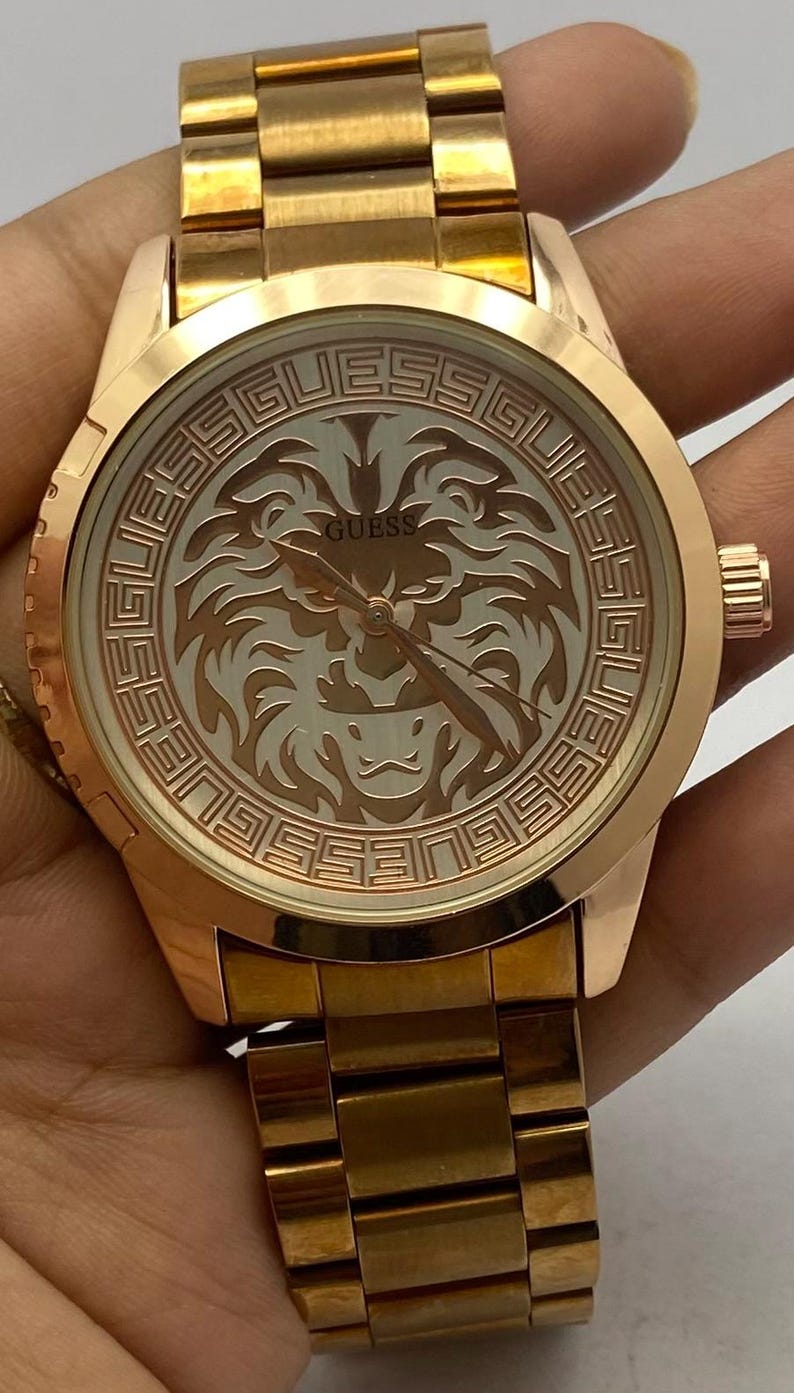 May include: Rose gold-toned Guess watch with a round face. The watch face features a lion design and the Guess logo. The watch has a matching rose gold band. The watch is a stylish accessory.