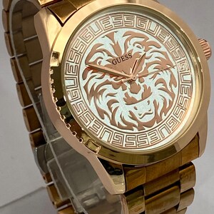 May include: Gold-toned Guess watch with a round face featuring a lion design. The watch has a gold band and a textured bezel. The watch face has the word "GUESS" and a lion graphic.